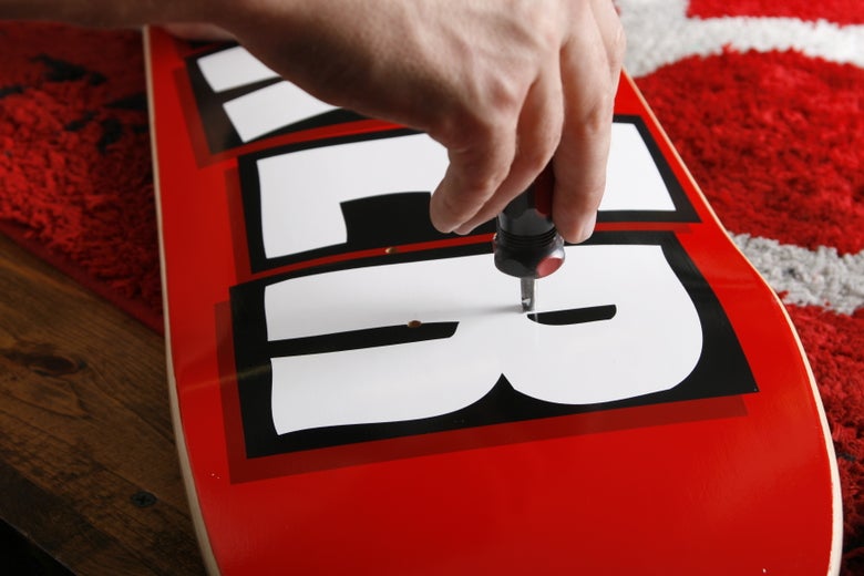 A person sticks a poker through the hardware holes of a skateboard deck to be able to insert hardware into the skateboard.