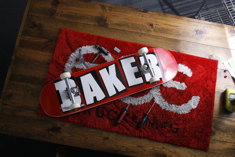 A complete Baker skateboard on a work bench with a skate tool, poker, power drill, razor blade, and skate components. 
