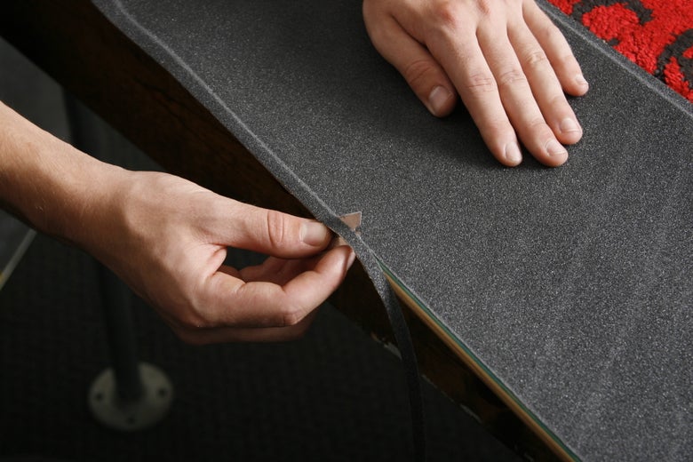 A person uses a razor blade to cut the excess griptape around the edges of the skateboard deck.