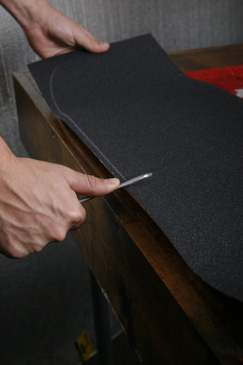 A person uses a screwdriver to scrape around the edges the skateboard deck to further apply the griptape and create cutting lines. 